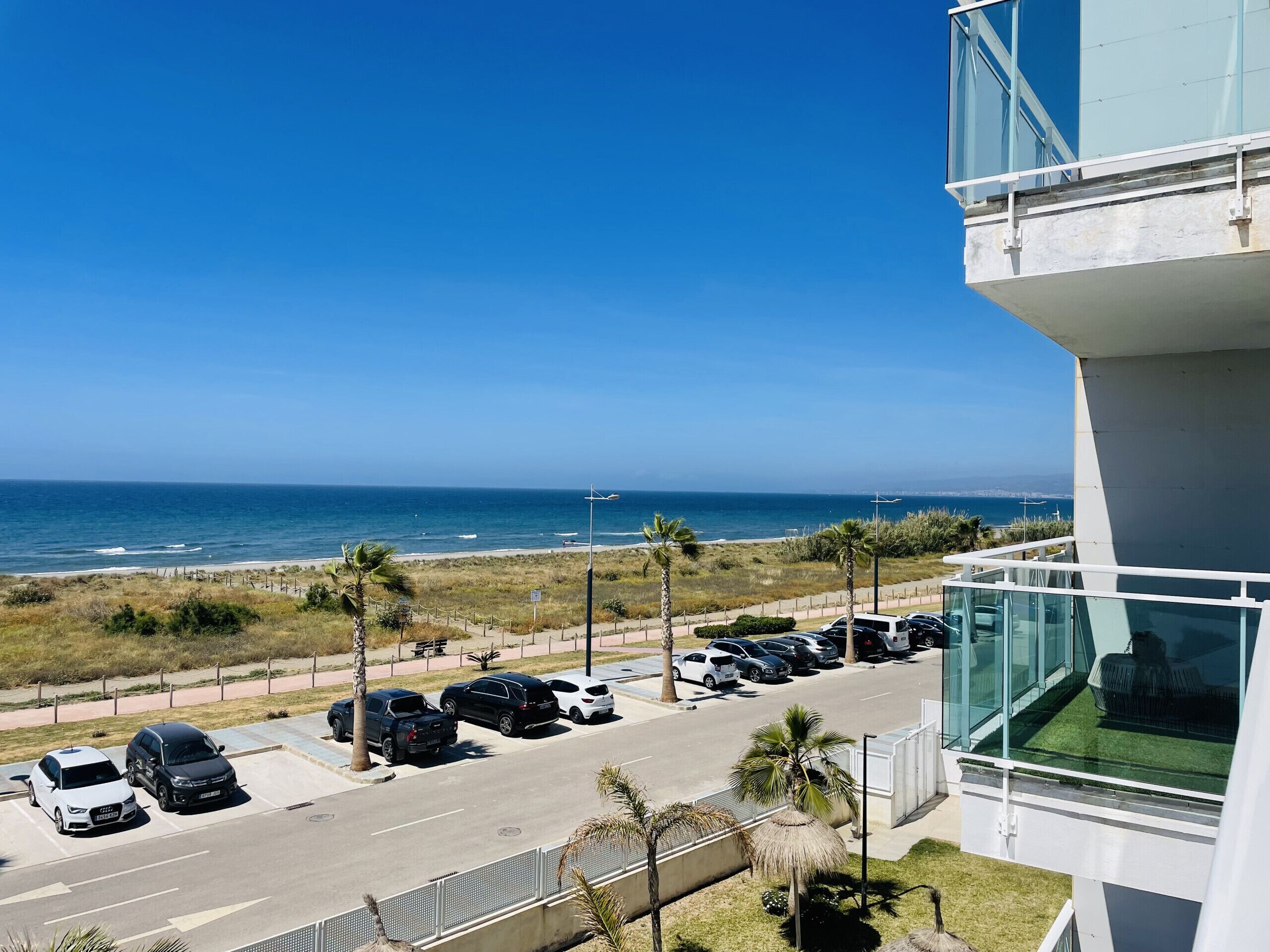 A parking area with several cars, a beach, and a clear blue sky.