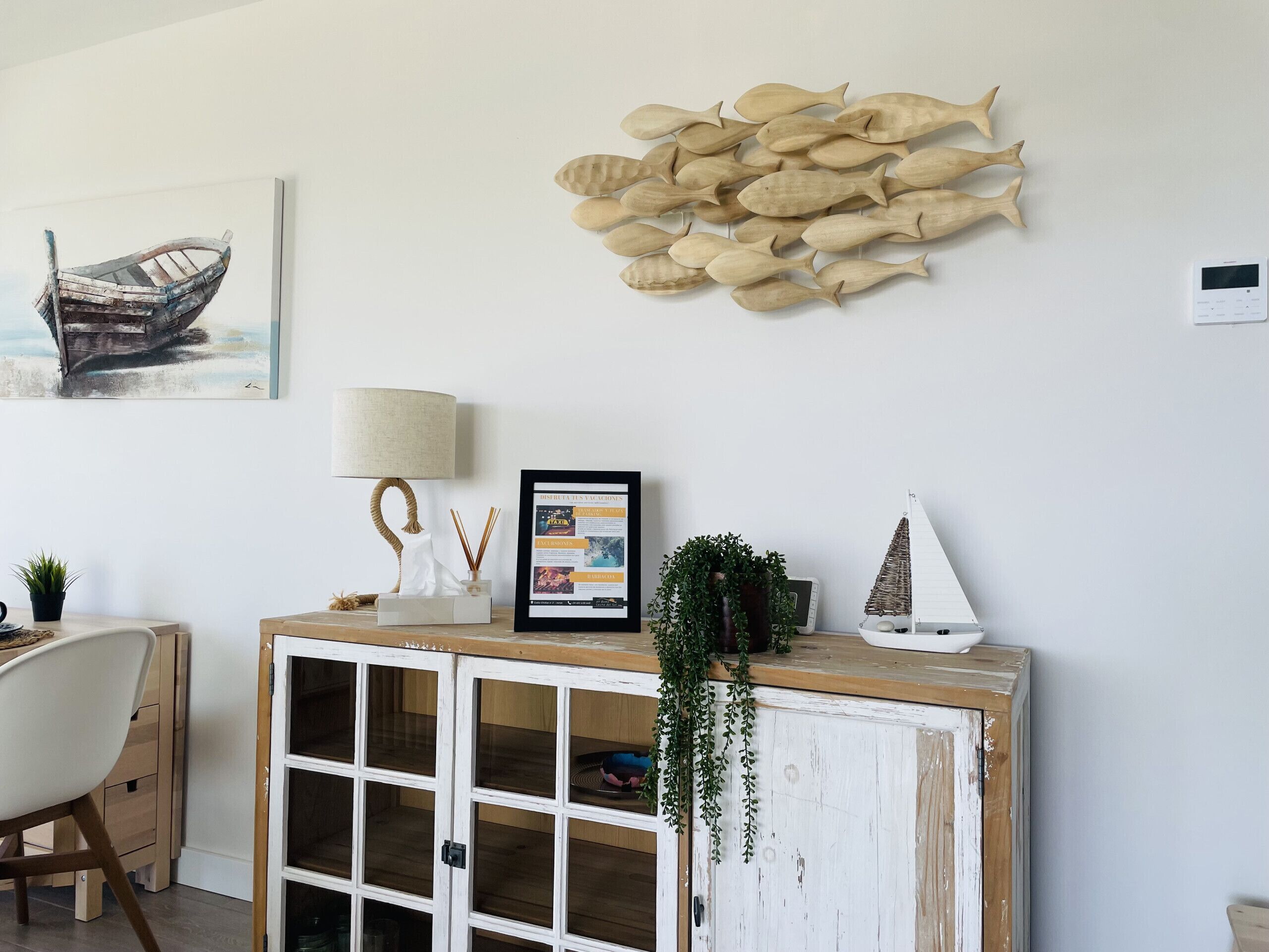 Een kamer met een witte muur, een lijst met een foto van een boot, een visbeeld aan de muur, een lamp, een menukaart in een lijst, een potplant en een kast met glazen deuren.