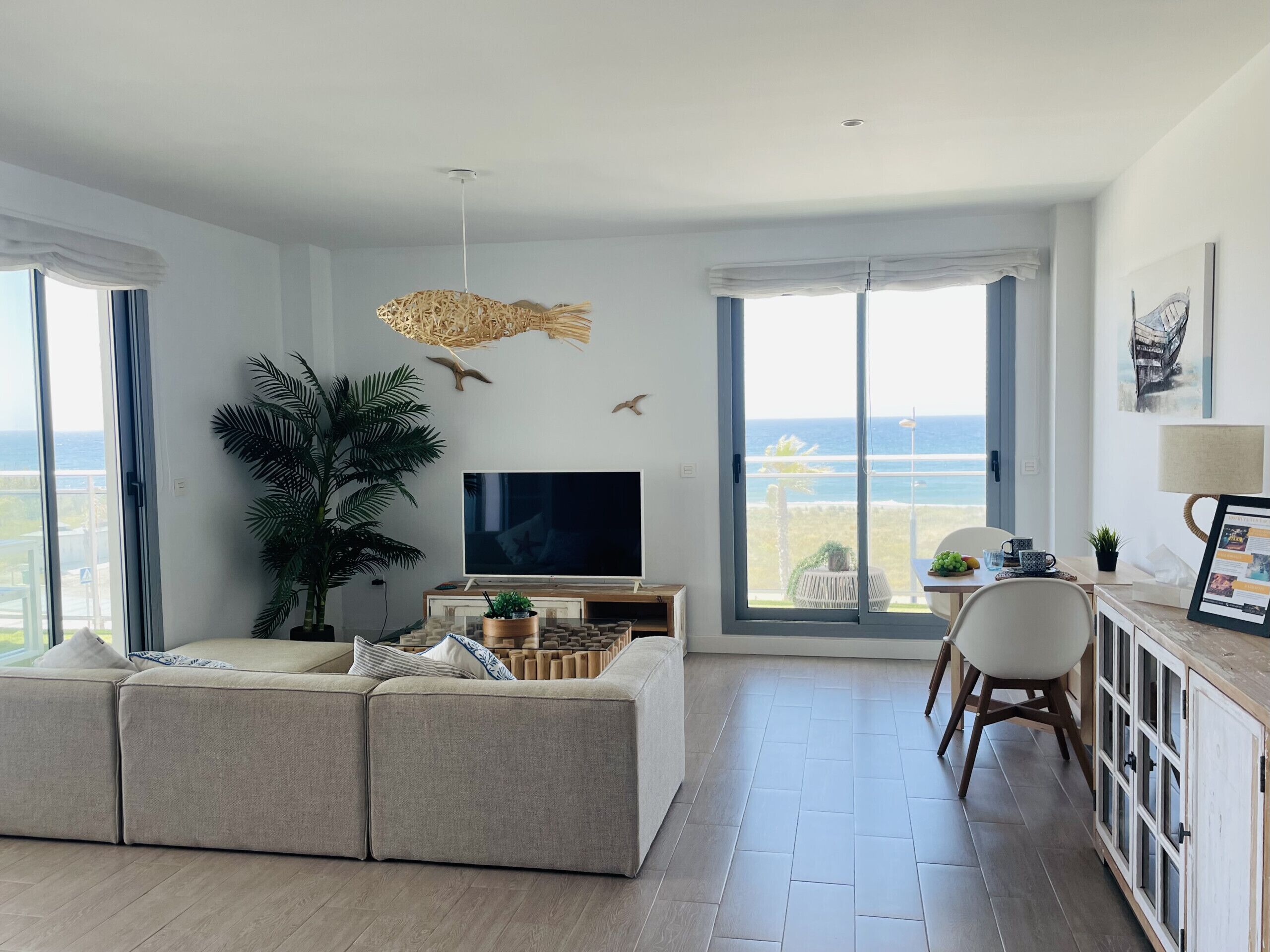 A modern living room with a large sofa, a flat-screen TV, a dining area with a table and chairs, and a view of the sea through large windows.
