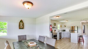 Un coin repas comprenant une table, des chaises et un chemin de table à motifs floraux. On aperçoit également une cuisine équipée avec des appareils électroménagers et des placards.