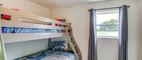 Une chambre avec un lit superposé, un ventilateur de plafond, une fenêtre avec des rideaux et une vue sur la verdure à l’extérieur.