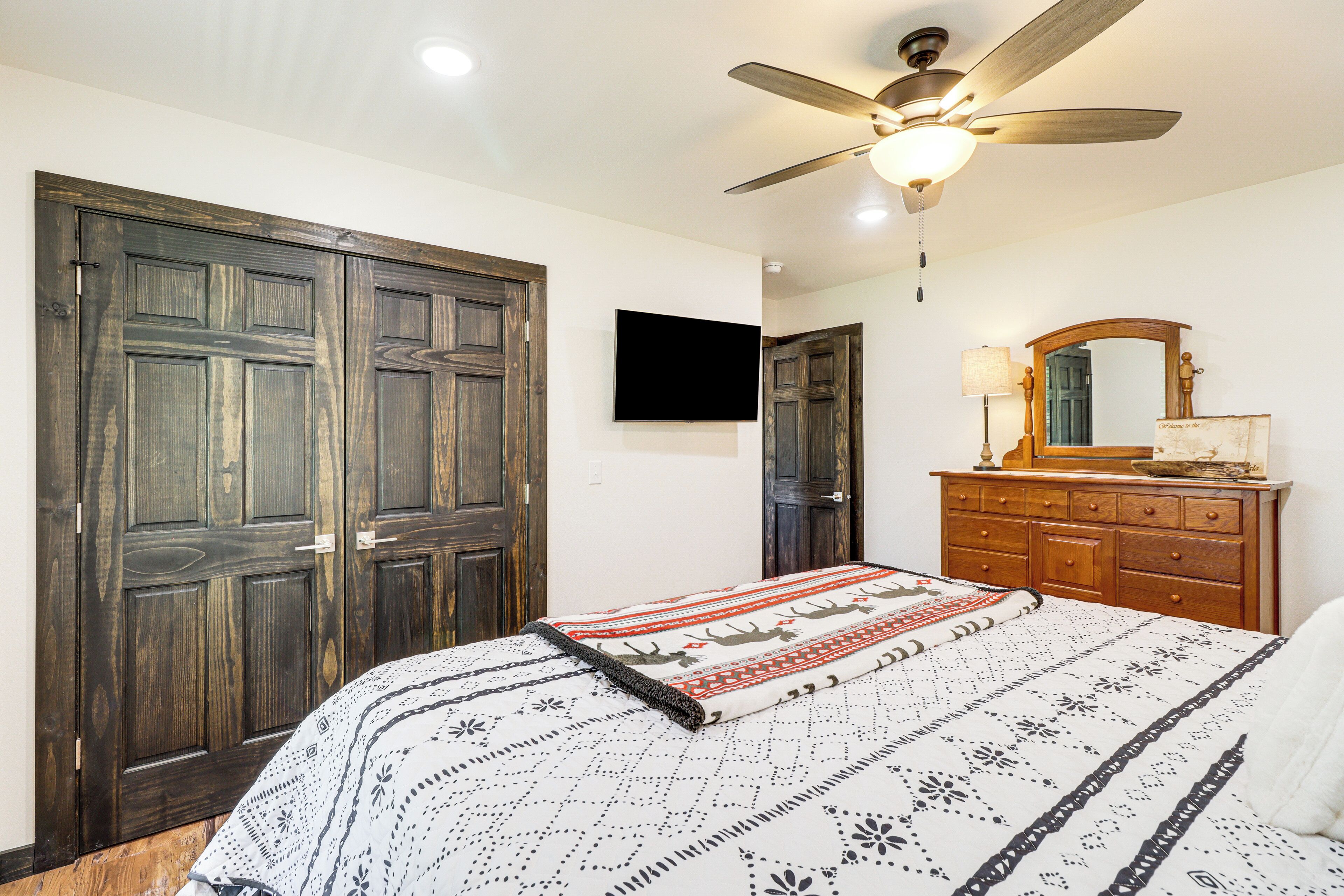Une chambre à coucher comprenant un lit, un ventilateur de plafond, un téléviseur fixé au mur et une commode avec un miroir.