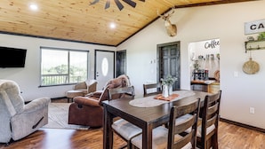 A spacious living room with a wooden ceiling, a dining area with a wooden table and chairs, a mounted deer head, and a coffee station.