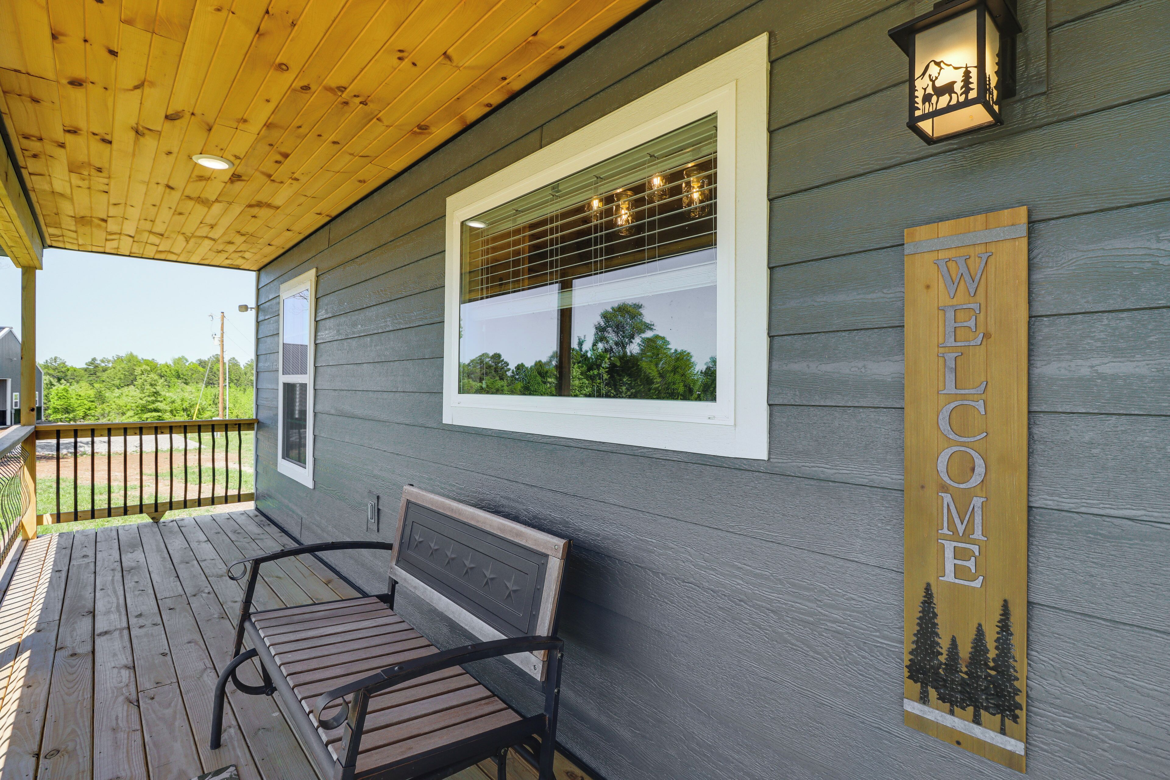 Una terraza de madera con un banco, un cartel de bienvenida y una ventana que da a un paisaje de árboles.