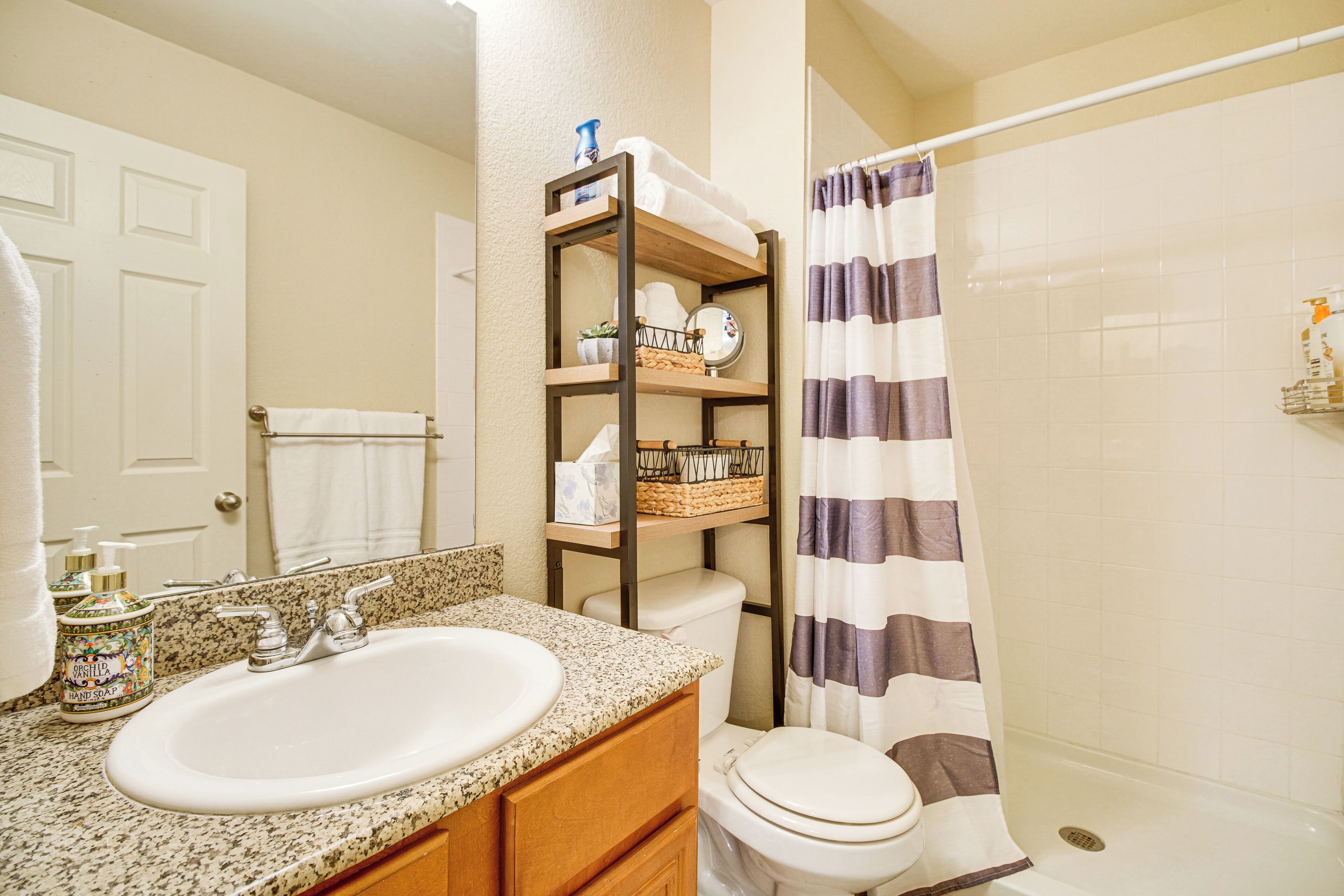 Une salle de bain avec un lavabo surmonté d’un plan de travail en granit, une étagère en bois où sont rangés les articles de toilette, des toilettes blanches et un rideau de douche.
