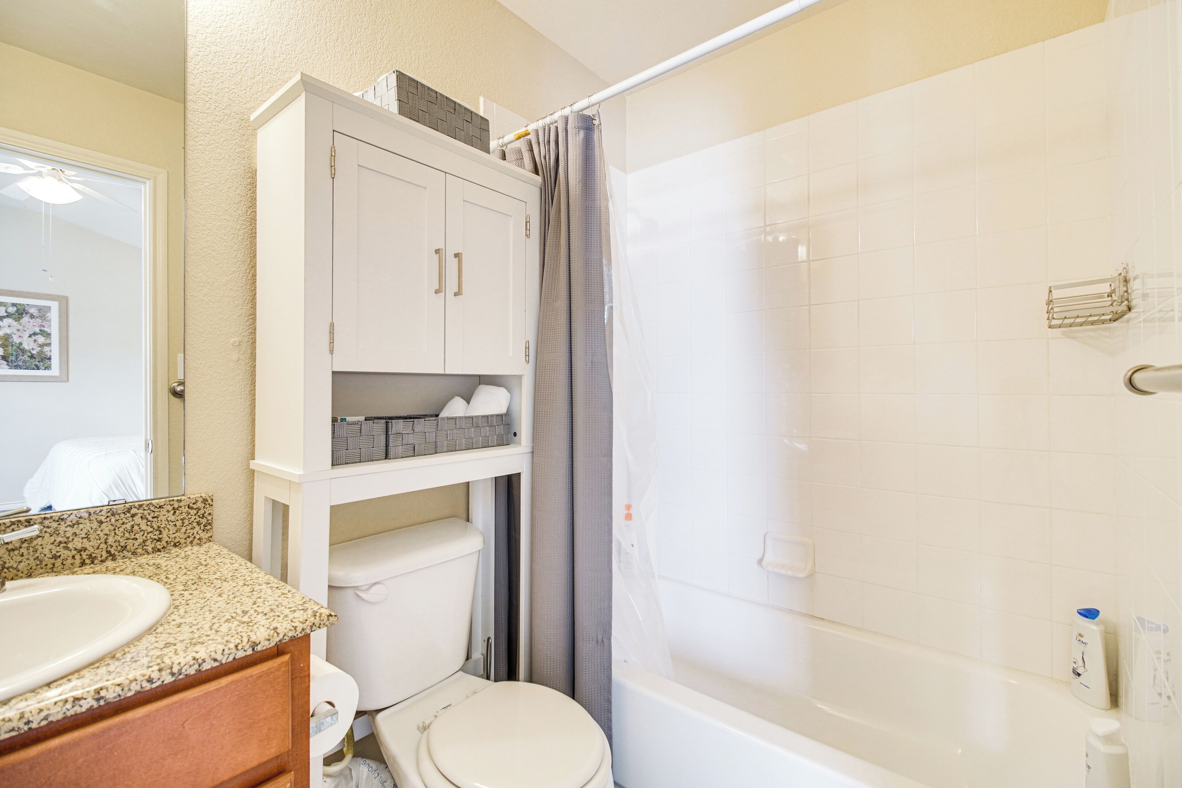 Une salle de bain avec des toilettes blanches, un lavabo doté d’un plan de travail en granit, une douche avec une porte en verre et un meuble blanc au-dessus des toilettes.