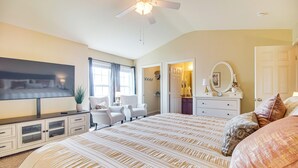 Une chambre avec un grand lit, un ventilateur de plafond, une télévision, une commode, une chaise et une porte donnant sur une salle de bain.