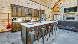 A modern kitchen with wooden cabinets, a central island with bar stools, a built-in oven, and a stone fireplace. - Private Hot Tub: Luxe Cabin in Broken Bow! (Broken Bow)