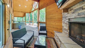 A wooden cabin with a stone fireplace, wicker furniture, and a large TV screen. - Private Hot Tub: Luxe Cabin in Broken Bow! (Broken Bow)