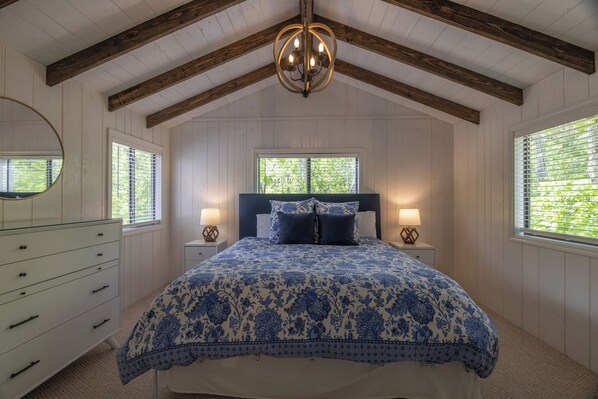 A bedroom with a large bed, wooden beams, a chandelier, two bedside tables with lamps, a dresser, and windows with blinds.