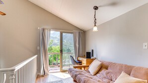 A cozy living room with a sofa, a desk, and a chair. There's a large window with curtains, and a balcony is visible. - Beautiful Kernville Home w/ Game Room & Mtn Views! (Kernville)
