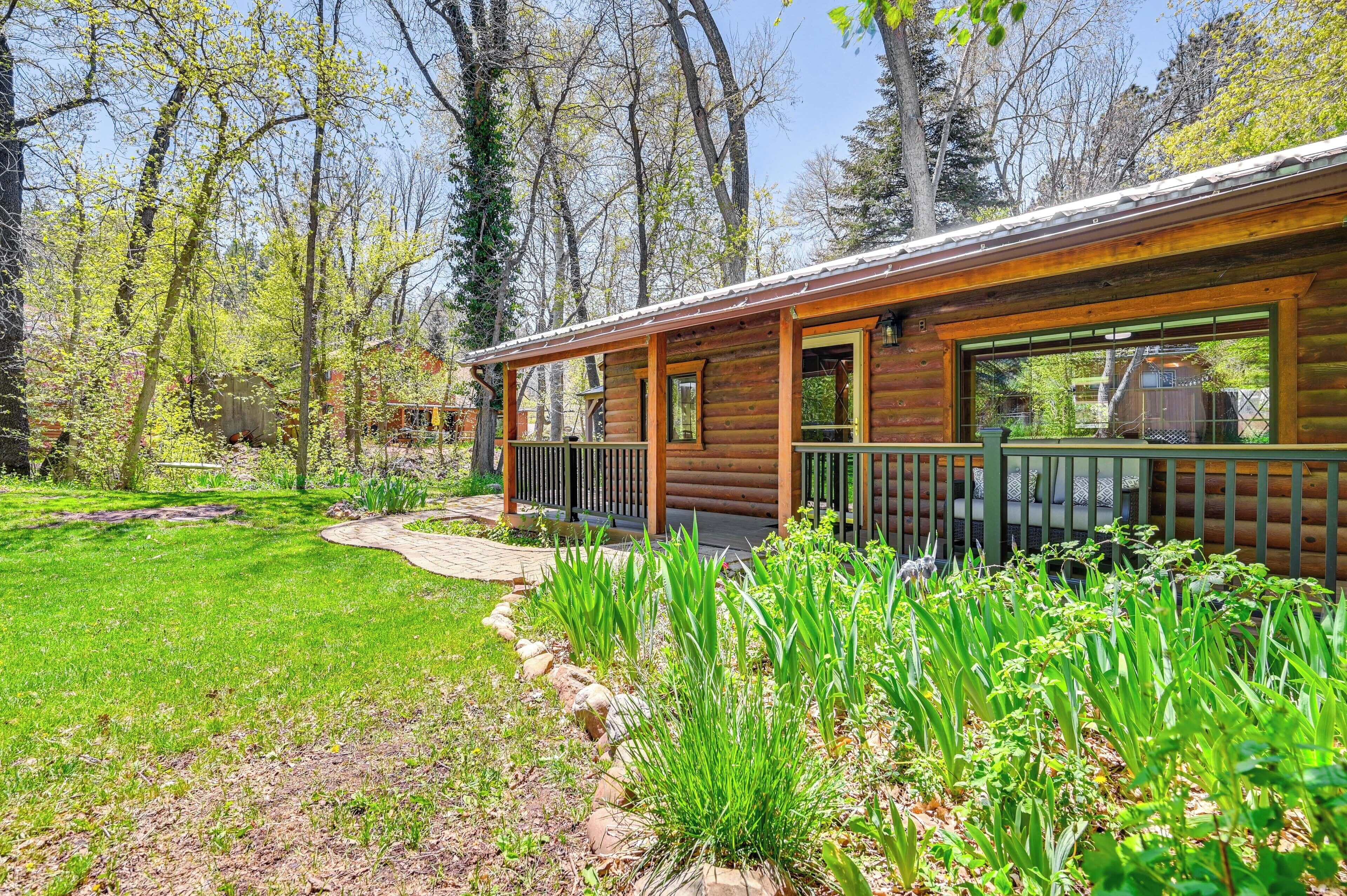 A log cabin with a porch, surrounded by a well-maintained garden and tall trees.
