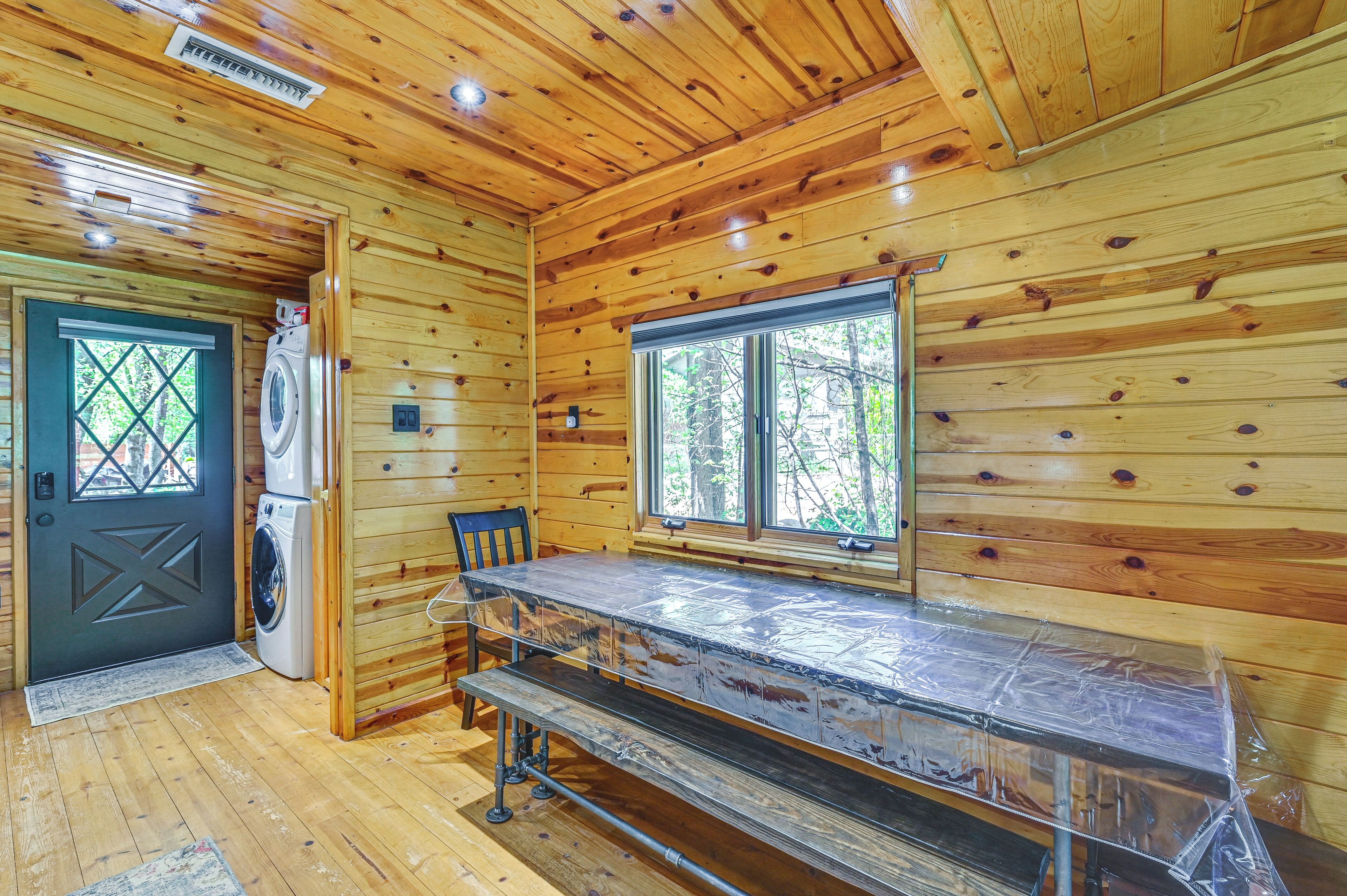A wooden cabin interior with a washing machine, a bench, and a window with a view of trees.