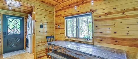 A wooden cabin interior with a washing machine, a bench, and a window with a view of trees.