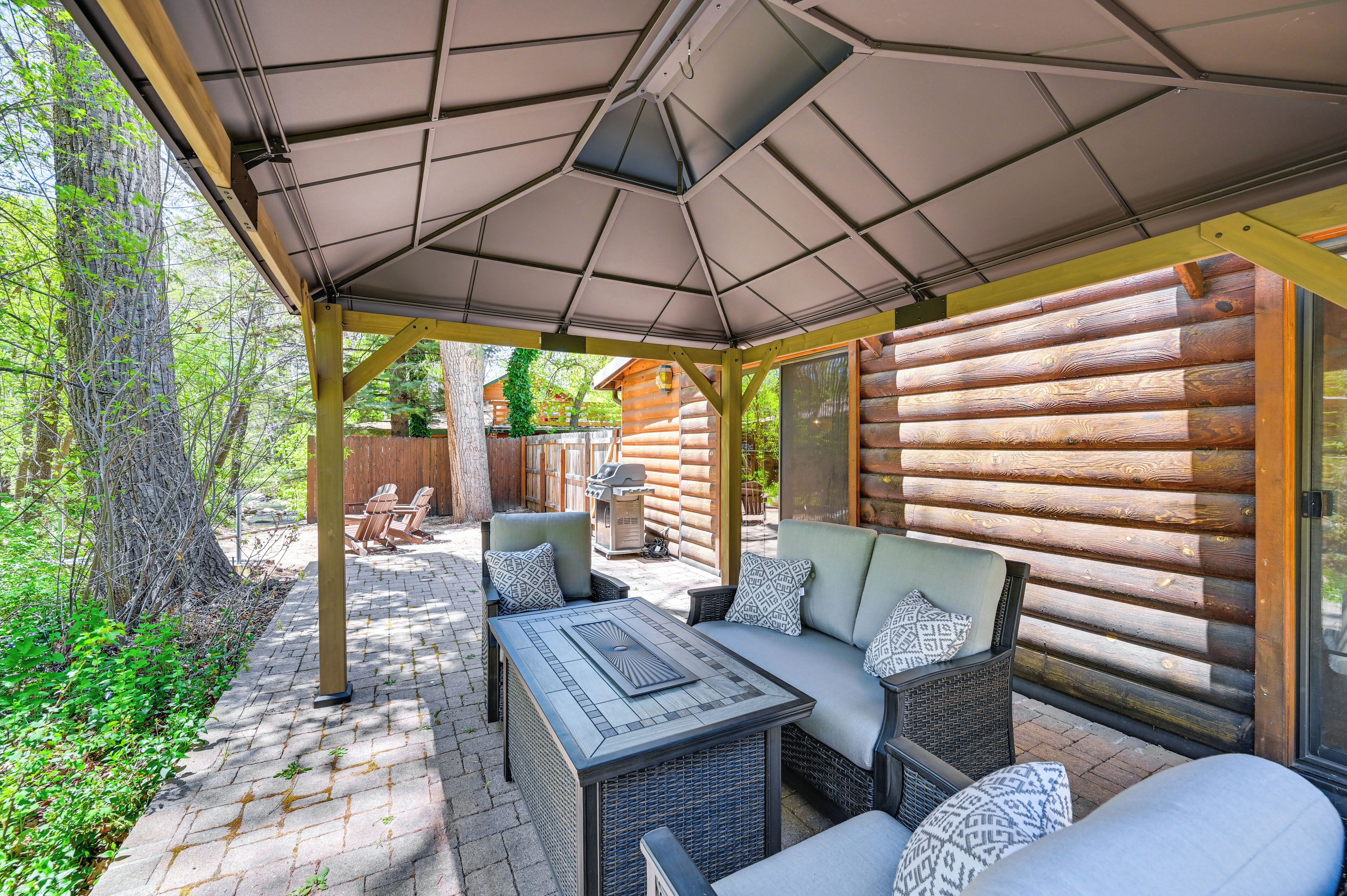 A covered patio with a table and seating, surrounded by wooden log walls and a view of a garden.