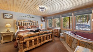 A rustic bedroom with a wooden bed, a stone bathtub, and large windows offering a view of a snowy landscape. - Quiet Fall River Escape • River Views • RMNP (Estes Park)