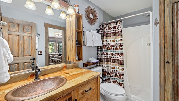 A bathroom with a wooden vanity, a large mirror, a white toilet, and a shower curtain with animal patterns. - Quiet Fall River Escape • River Views • RMNP (Estes Park)
