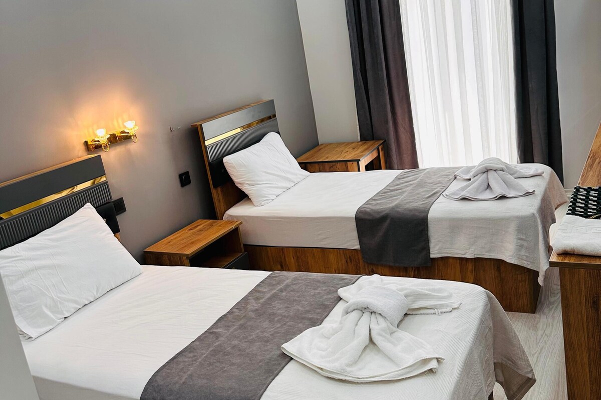 Habitación de hotel con dos camas, cabecera de madera, una mesita pequeña y una lámpara fijada a la pared.