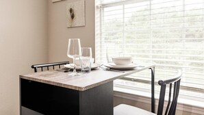 Una mesa de comedor con dos copas de vino, un bol y cubiertos. La mesa está cerca de una ventana con persianas, y hay dos obras de arte colgadas en la pared encima.