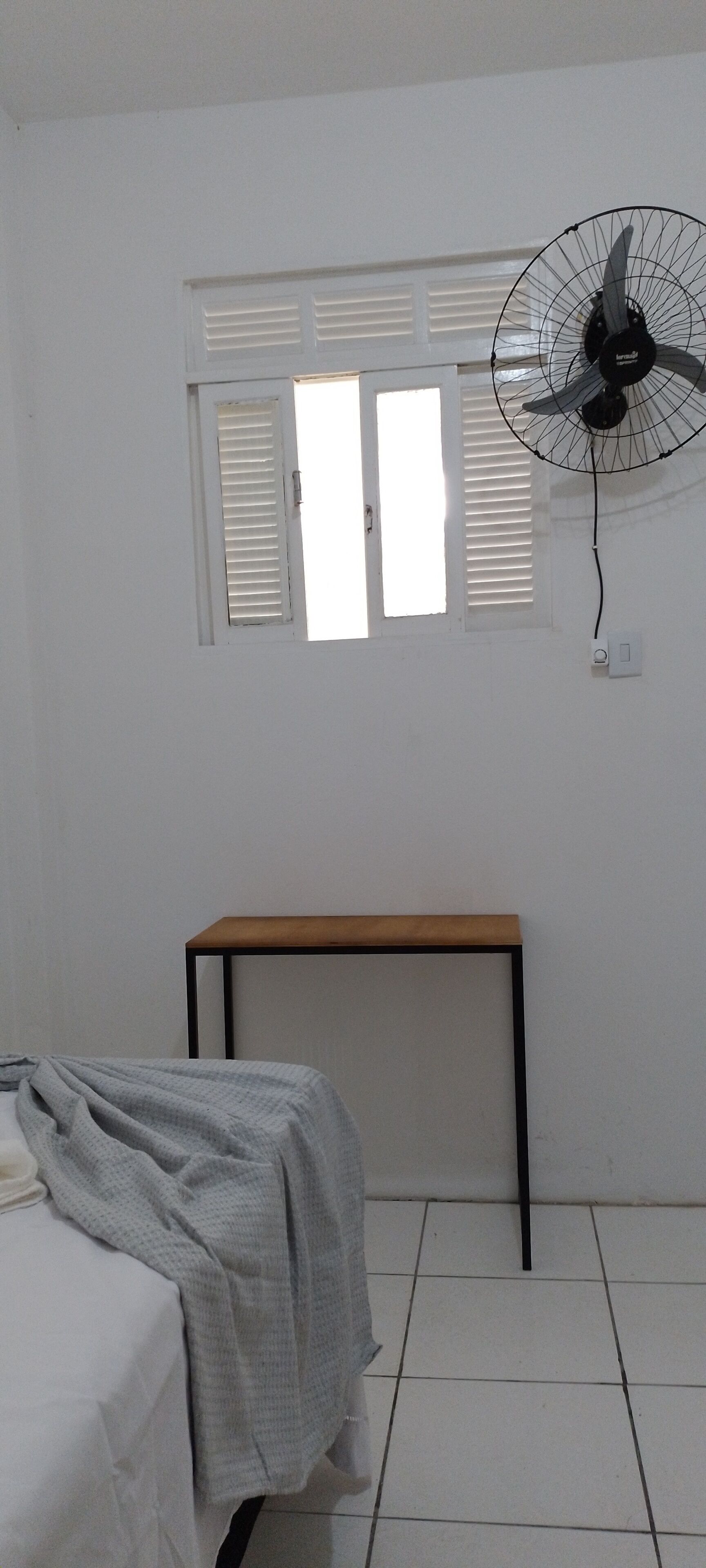 Una habitación con una cama, una mesa, un ventilador y una ventana con persianas.