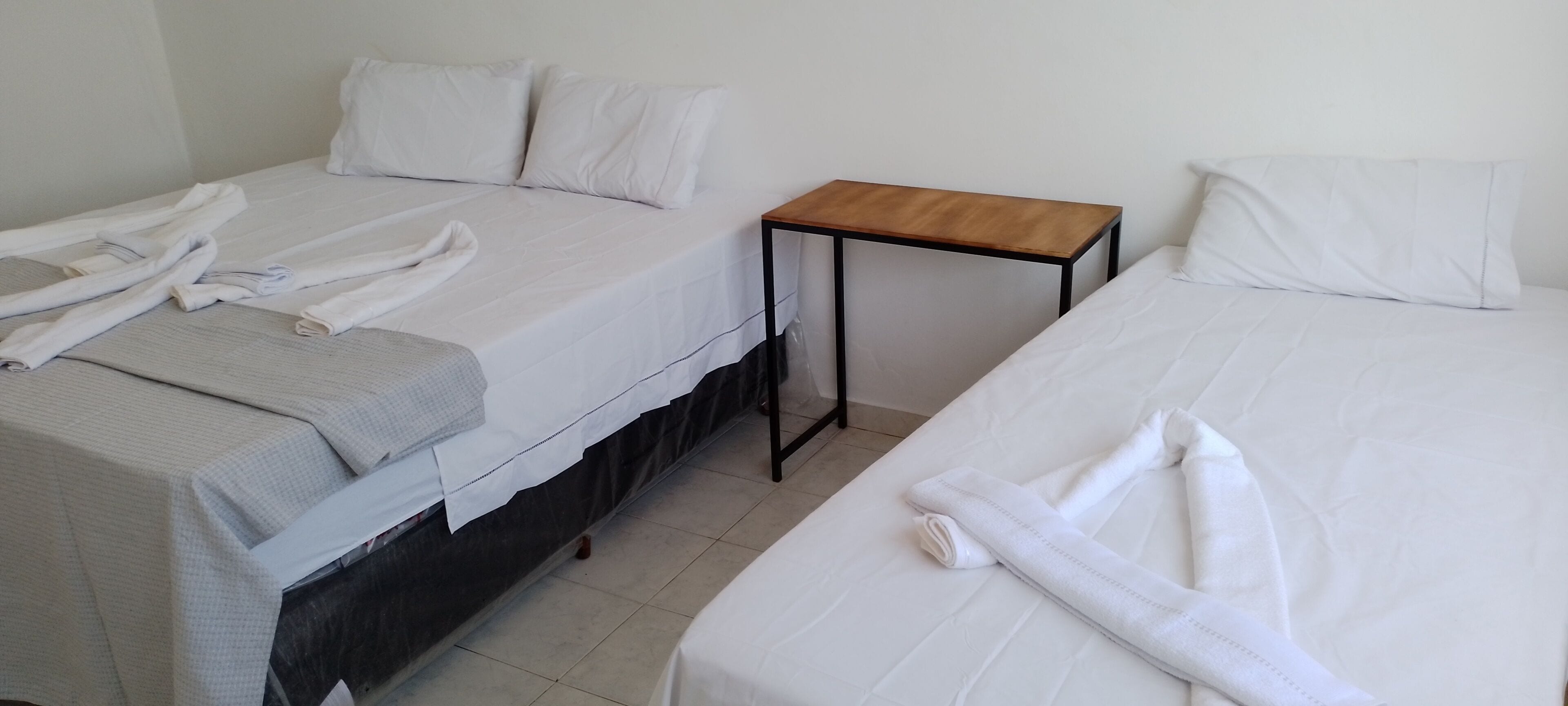 Habitación de hotel con dos camas, cada una con sábanas y almohadas blancas, una mesita de noche de madera y piso de baldosas.