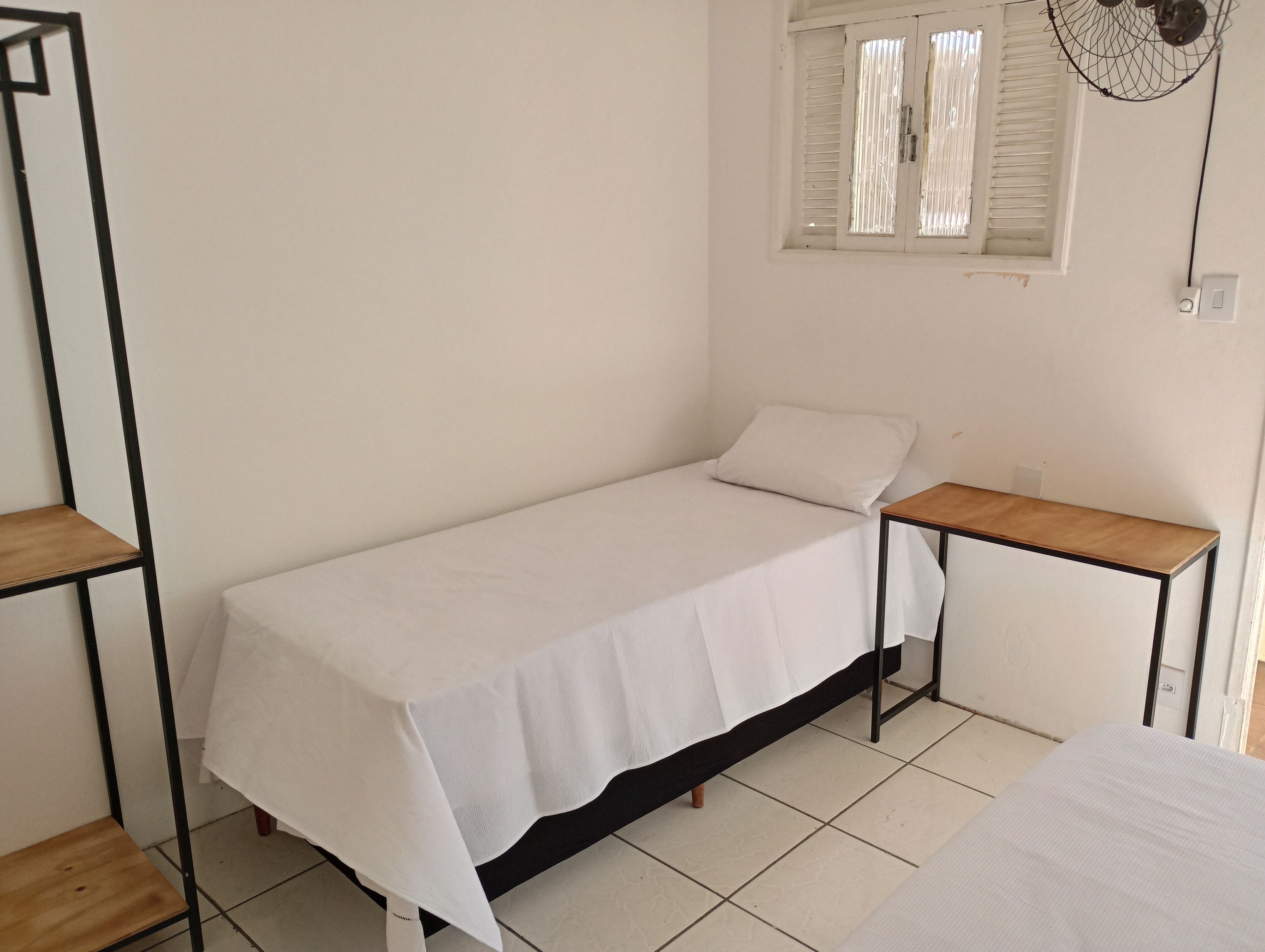 Una habitación pequeña con una cama individual, una mesita de noche de madera, una ventana con persianas y un ventilador fijado a la pared.