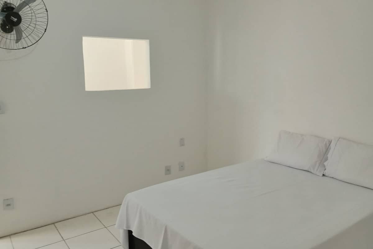 Una habitació amb un llit, un ventilador fixat a la paret i una finestra petita.