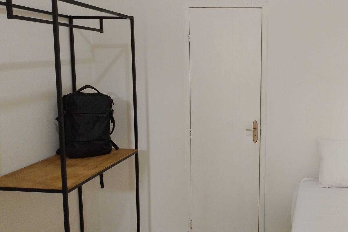 Una habitació amb un llit, una porta blanca i una motxilla negra sobre una prestatgeria.