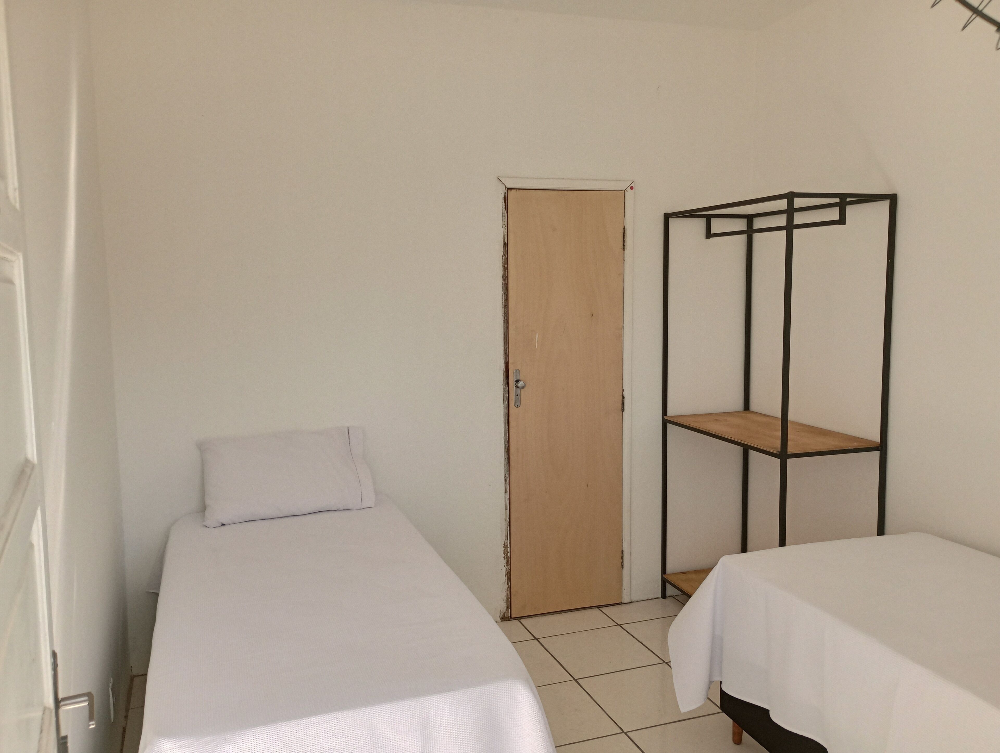 Una habitación pequeña y escasamente amoblada con dos camas individuales, una puerta de madera y un estante metálico.