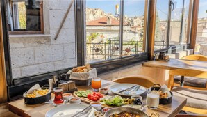 A dining table with various dishes, including a pan of food, plates, and bowls, in a room with a view of buildings through large windows.