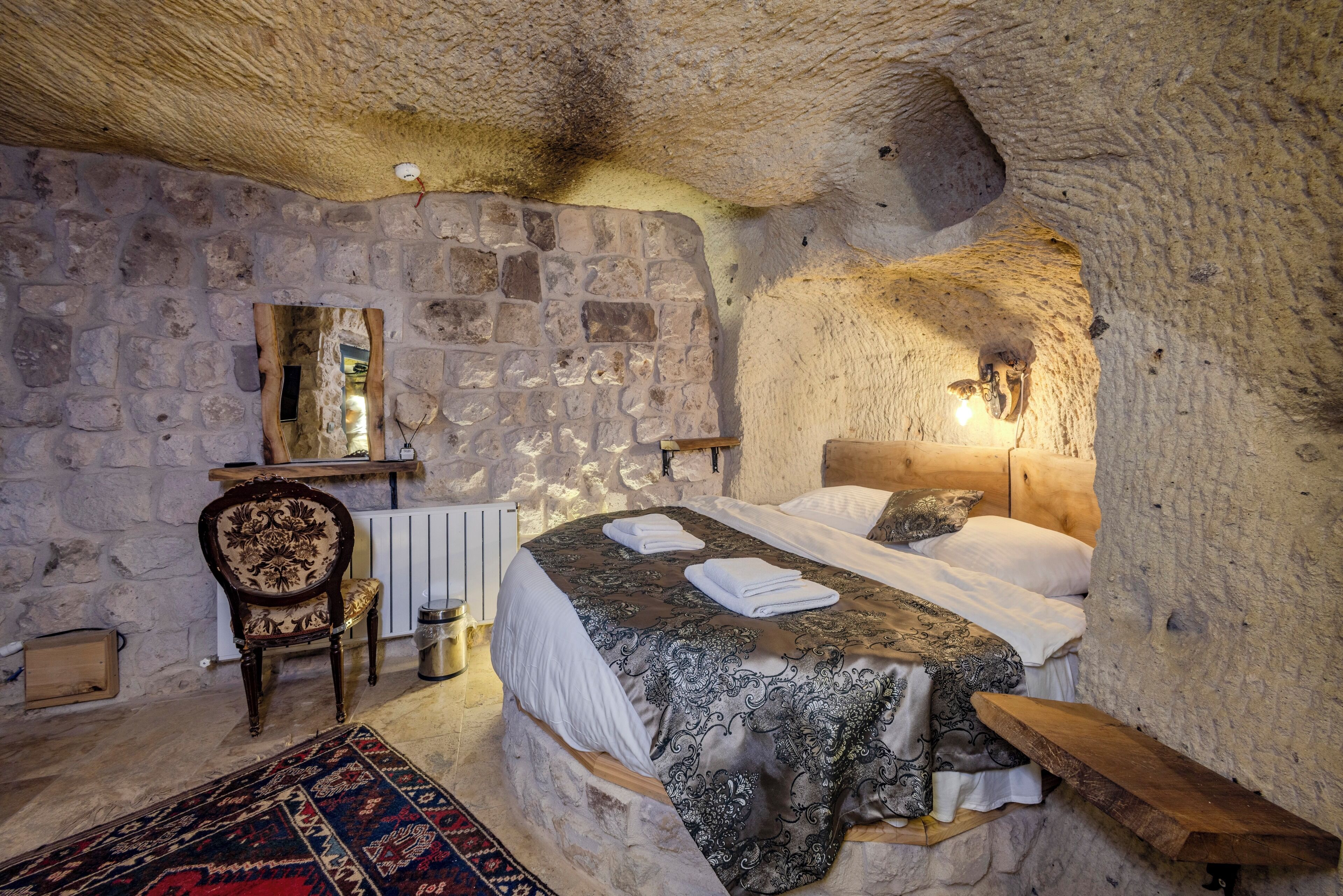 A stone-walled bedroom with a bed, a wooden chair, a radiator, and a mirror on the wall.