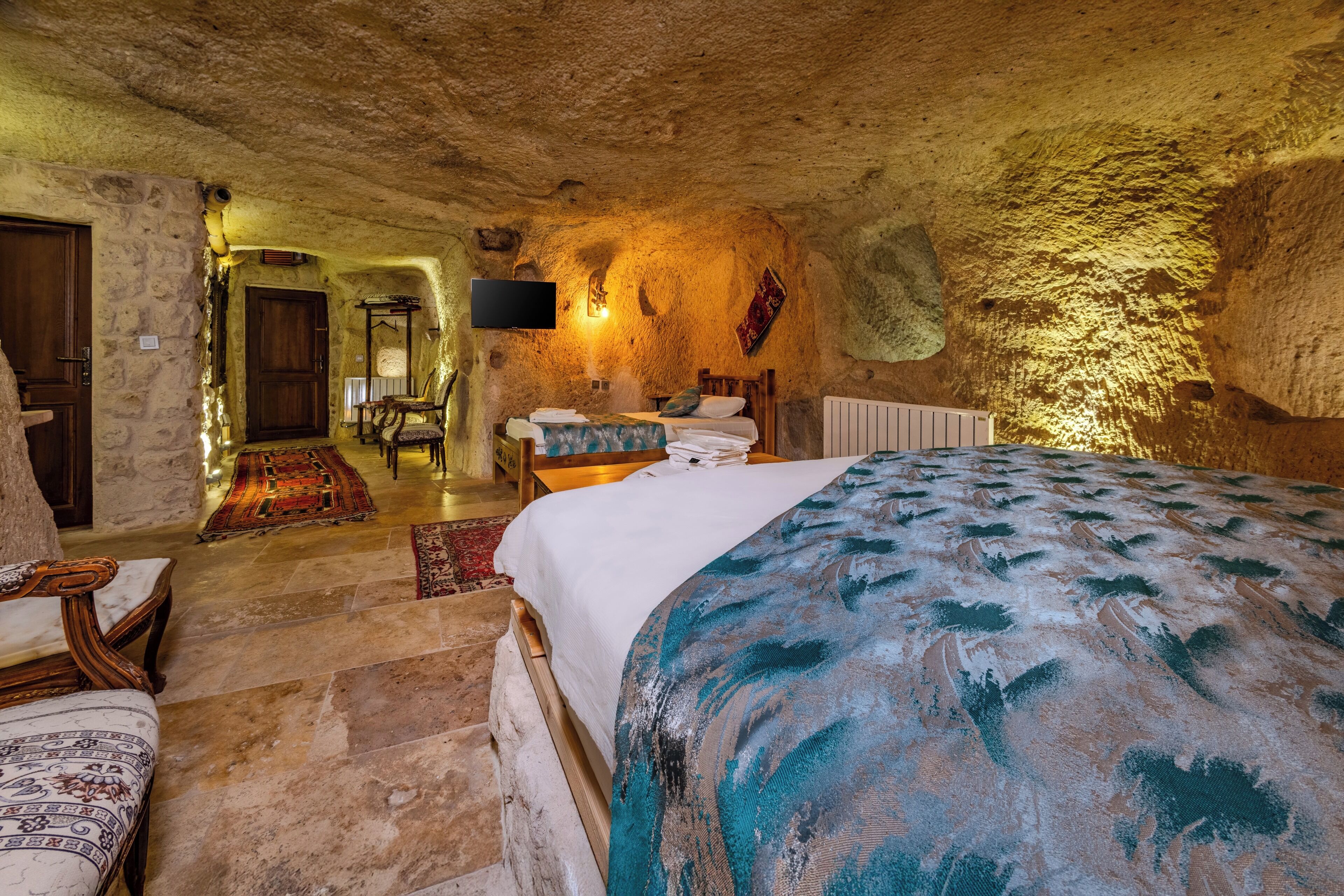 A rustic stone-walled bedroom with a bed, a wooden chair, a television, and a carpeted floor.