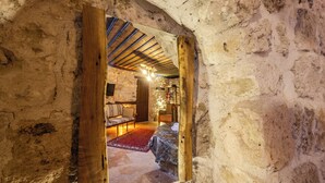 A rustic stone room with a wooden door leading to a cozy interior featuring a bed, a red rug, and a wooden ceiling.