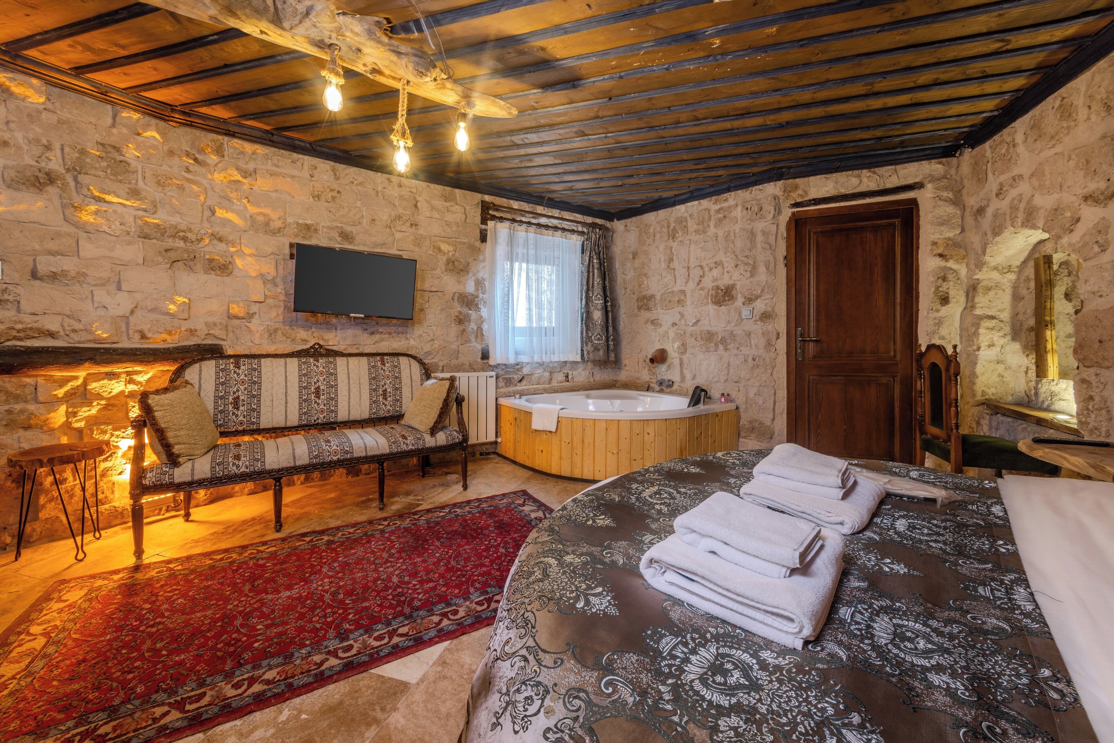 A rustic room with a stone wall, wooden ceiling beams, a large bed with a patterned bedspread, a wooden bench with cushions, a red rug, a wooden bathtub, and a wooden door.