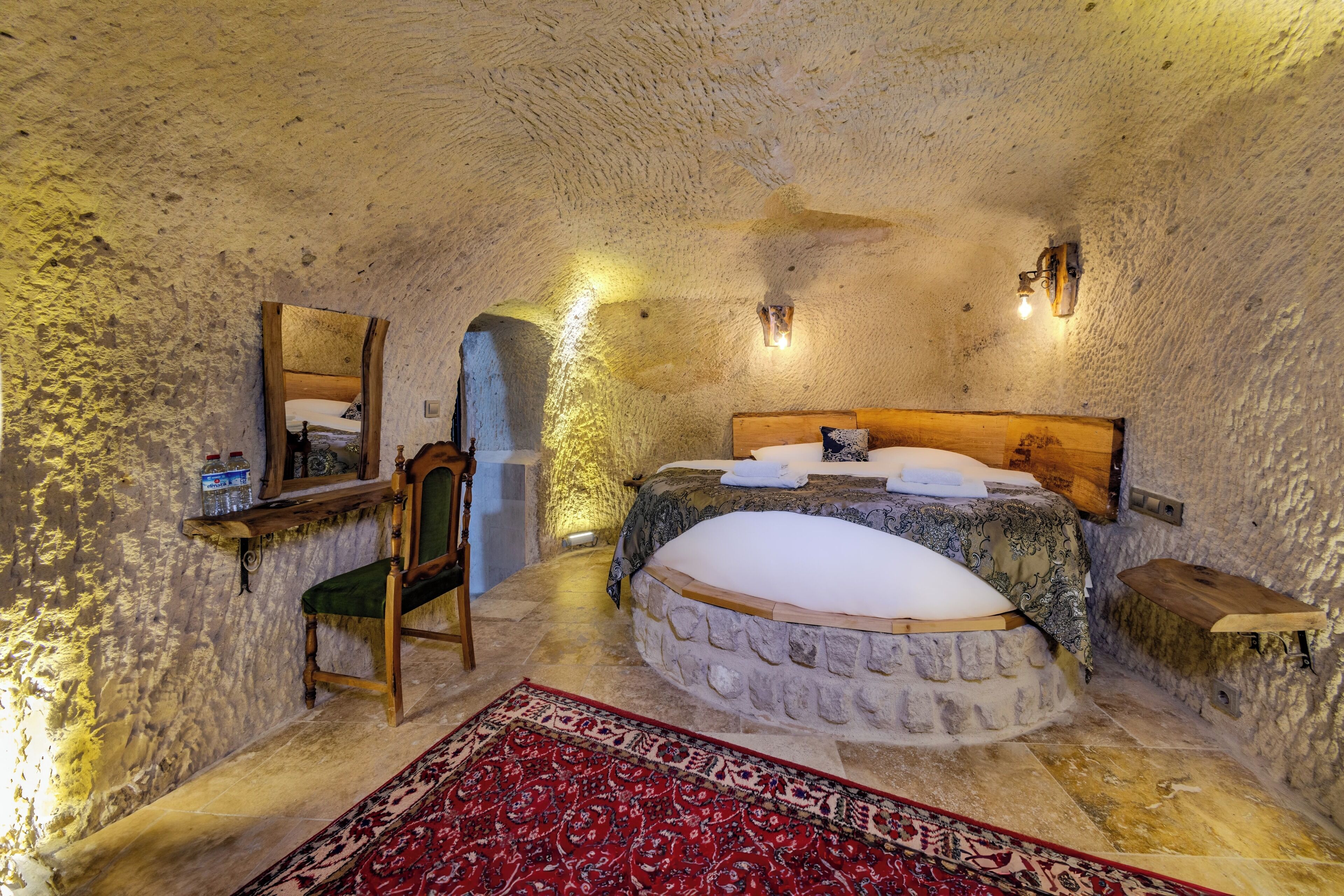 A rustic bedroom with a stone wall, a wooden bed, a chair, a mirror, and a red patterned rug.