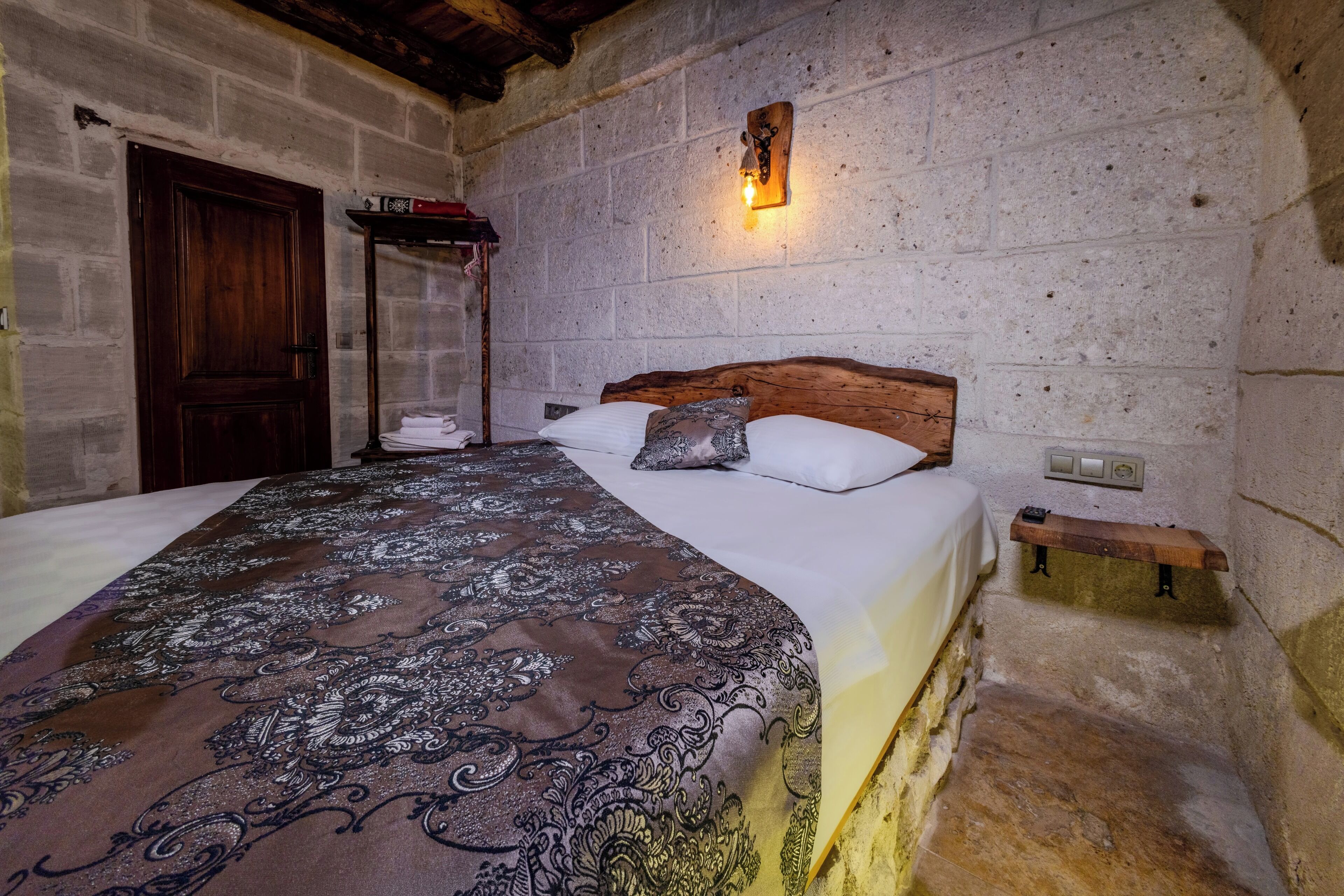 A bedroom with a large bed, a wooden headboard, a stone wall, a small shelf, and a wooden door.