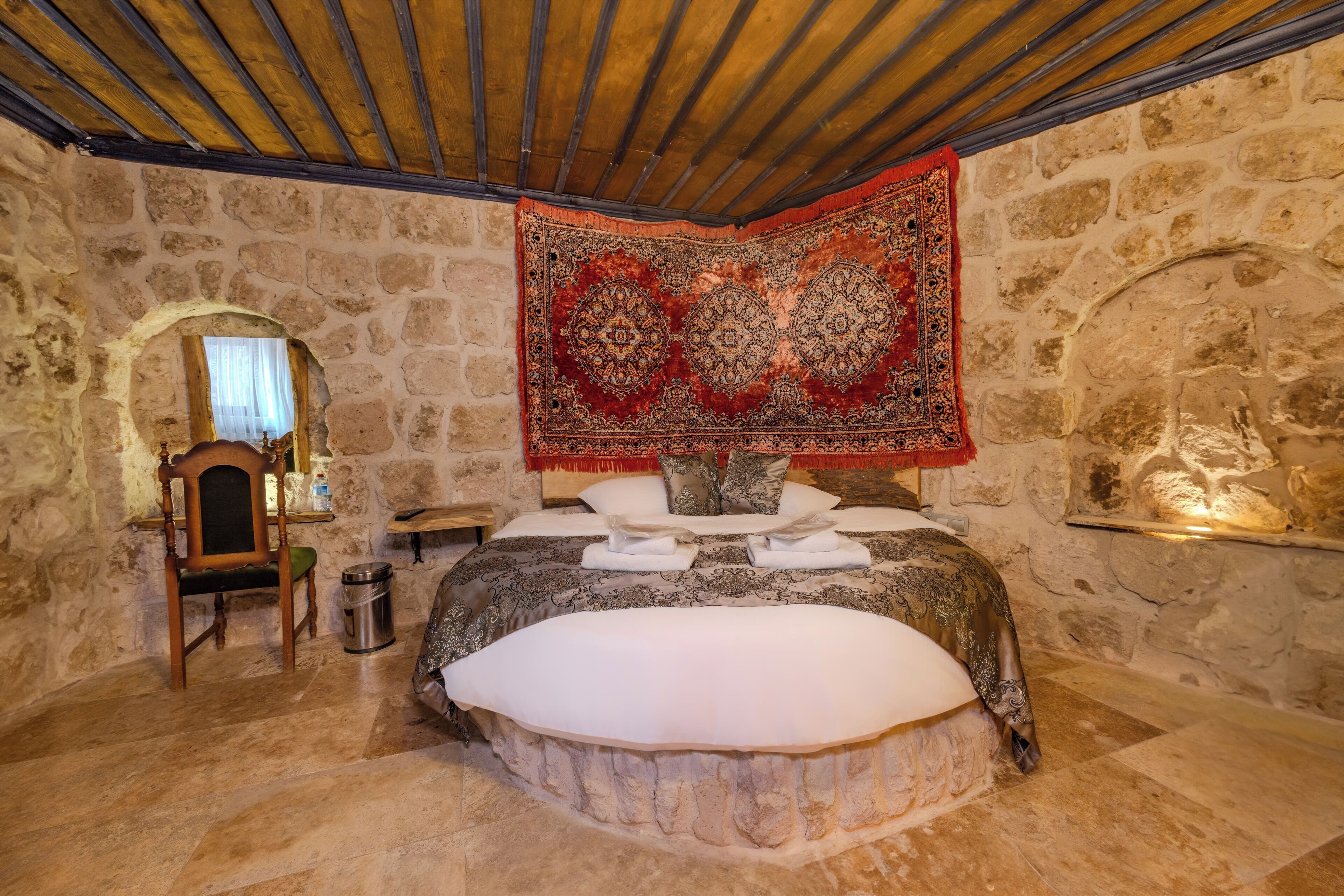Une chambre aux murs de pierre, avec un grand lit, une chaise en bois et une tapisserie rouge à motifs.