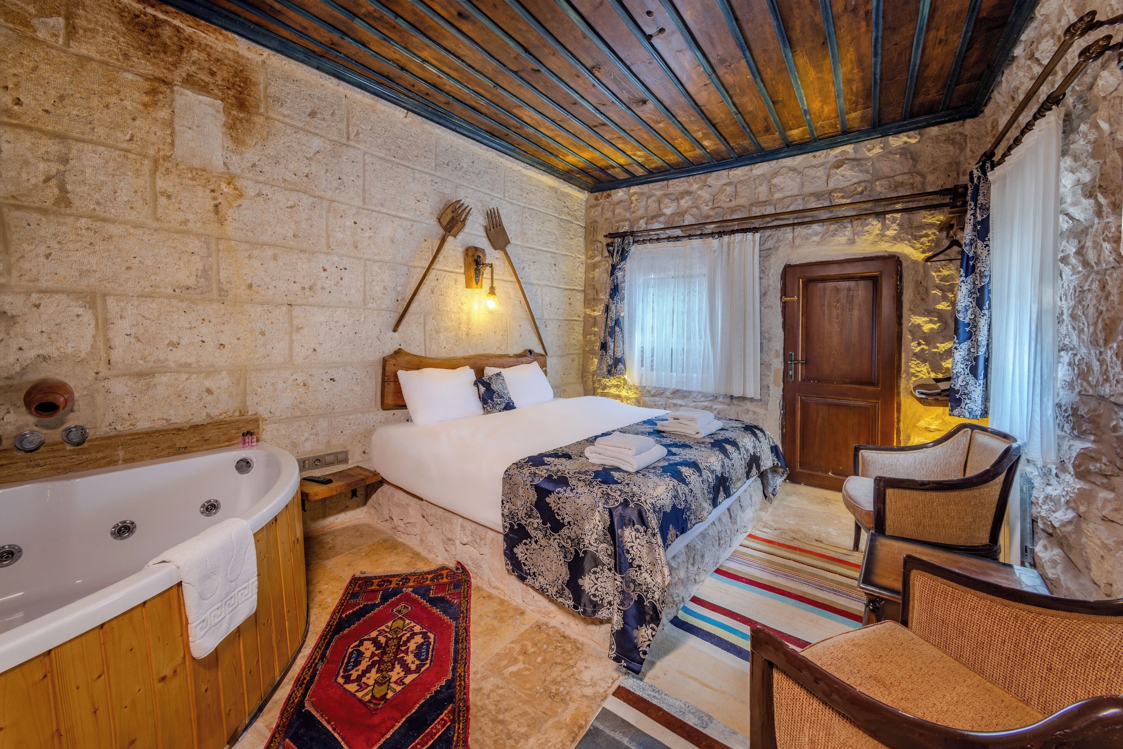 Een kamer met een stenen muur, houten plafond, een groot bed met een beddegoed met dessin, een houten stoel, een klein tafeltje en een bad met een handdoek.