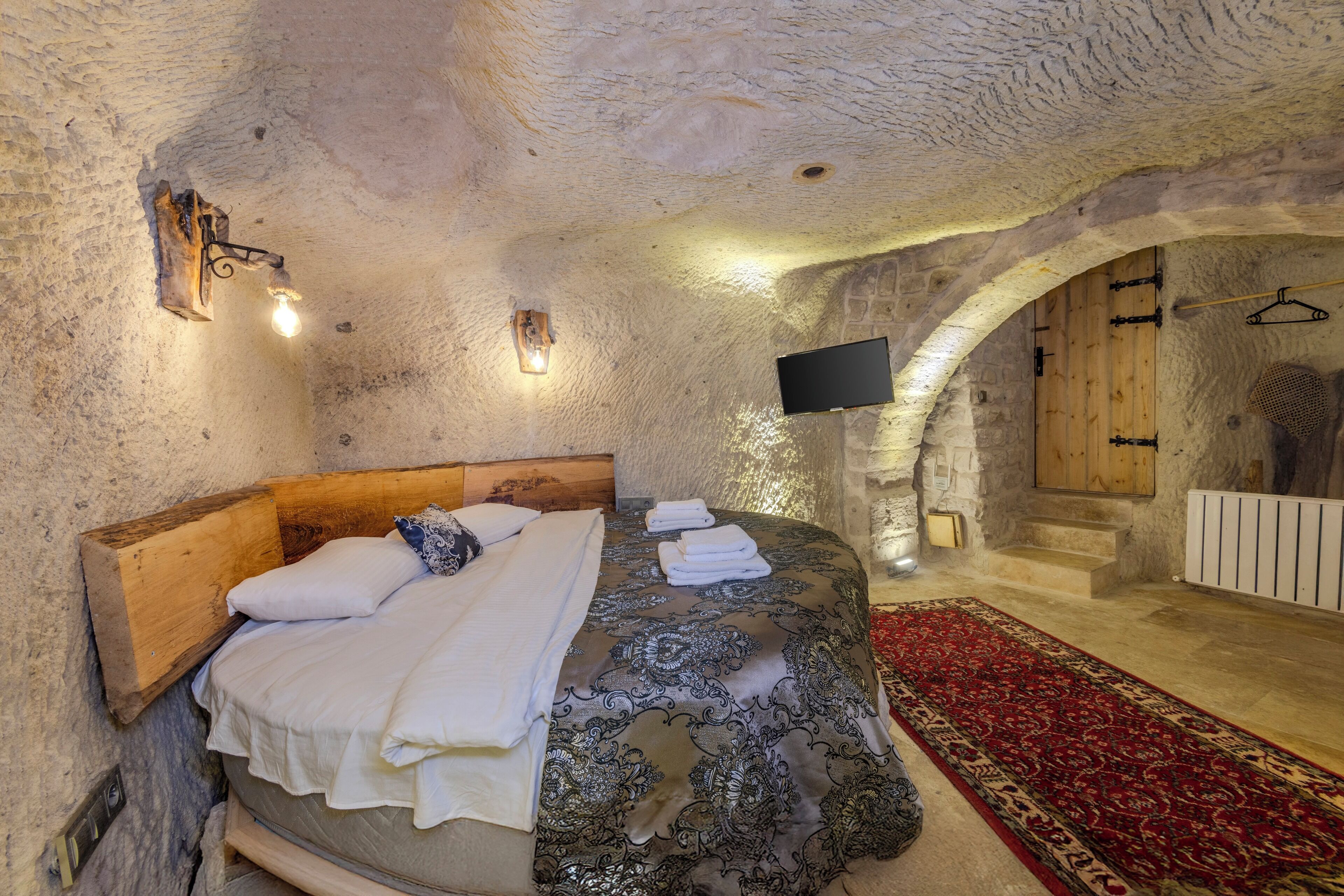 A bedroom with a stone wall, a wooden headboard, a bed with white linens, a patterned bedspread, a red and beige rug, and a flat-screen TV mounted on the wall.