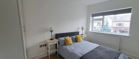 A bedroom with a bed, two bedside tables, a window, and a view of buildings outside.