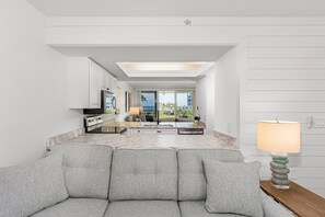 A modern living room with a grey sofa, a wooden side table with a lamp, and a view of a kitchen and balcony. - Oceanfront Condo in Seagate, the heart of Naples. (Naples)