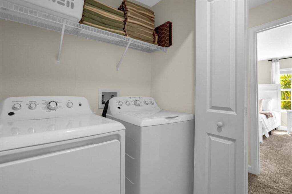 A laundry room with two stacked washers and dryers, a shelf above with folded linens, and a door leading to a bedroom with a bed and a view of greenery.