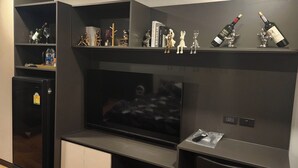 A modern living room with a flat-screen TV, a built-in mini-fridge, and a shelf with decorative items and wine bottles.