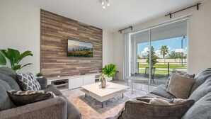 A modern living room with a flat-screen TV mounted on a wooden wall, a coffee table with a plant, and a large sliding glass door leading to an outdoor patio. - Last Minute Disney Getaway! Luxury Condo with Private Balcony. Book Now! #105363 (Orlando)
