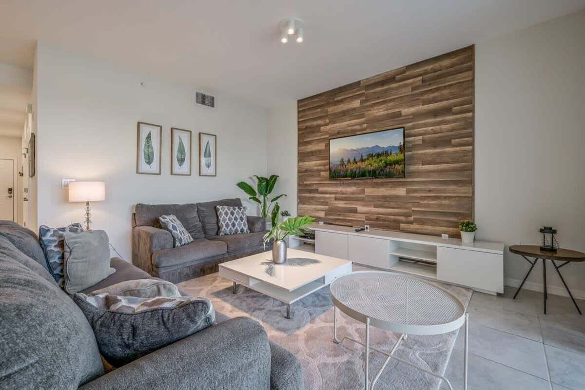Ein modernes Wohnzimmer mit einer grauen Couch, einem Couchtisch, einem an einer Holzwand montierten Fernseher und einem kleinen Beistelltisch.