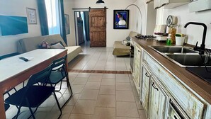 Une cuisine avec un plafond en bois, un coin repas avec une table et des chaises, et une vue sur le salon par une porte ouverte.