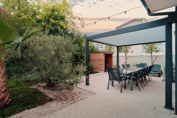 A covered patio area with a table and chairs, surrounded by greenery and a small wooden structure.
