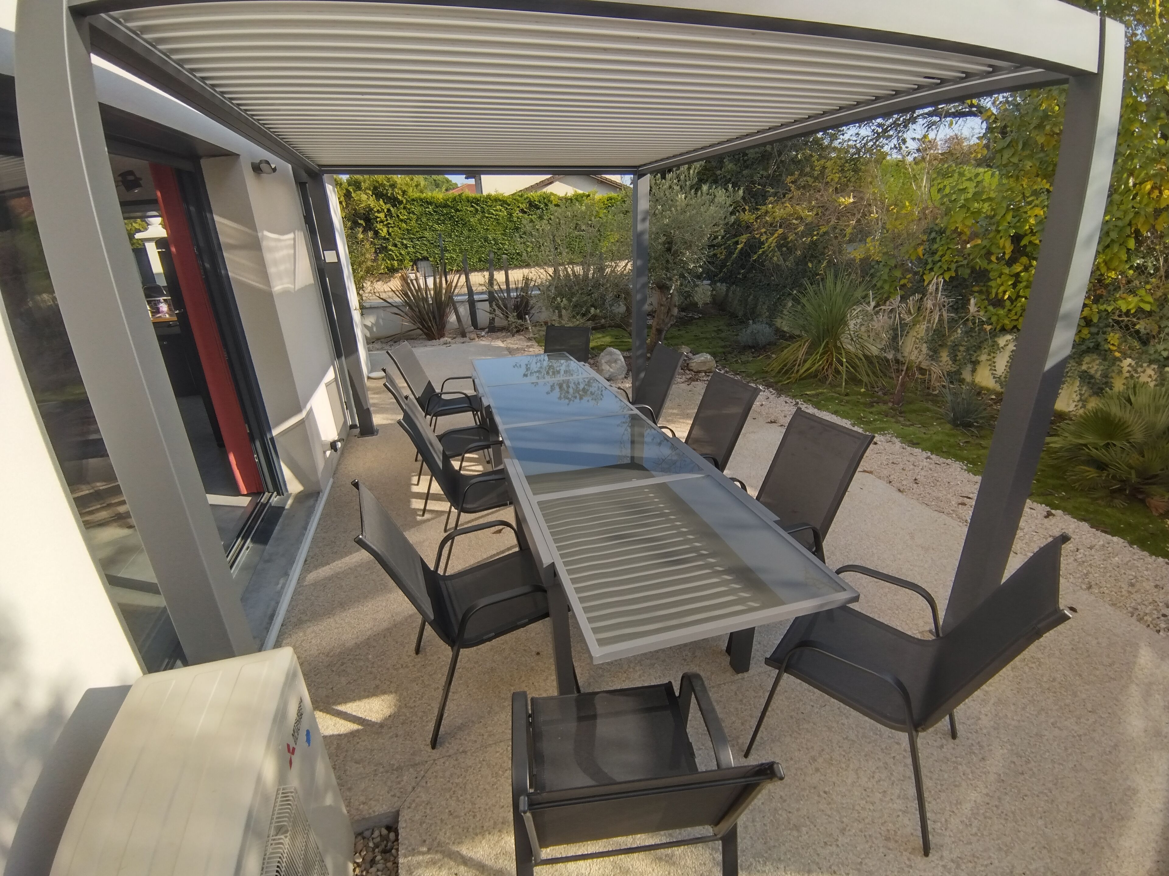 Un espace de restauration en plein air avec une longue table et des chaises, une pergola au-dessus et une vue sur un jardin où poussent différentes plantes.