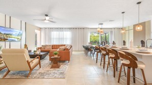 Ein modernes Wohnzimmer mit Sofa, Couchtisch und Sesseln, ein Essbereich mit Tisch und Stühlen sowie ein großes Fenster mit Vorhängen.