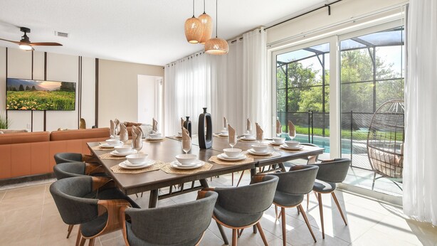 Ein moderner Essbereich mit einem langen Tisch, gedeckt für sechs Personen, umgeben von sechs grauen Stühlen mit Holzbeinen. Drei Pendelleuchten leuchten über dem Tisch, und ein großes Fenster bietet Blick auf einen Swimmingpool und Grünflächen.